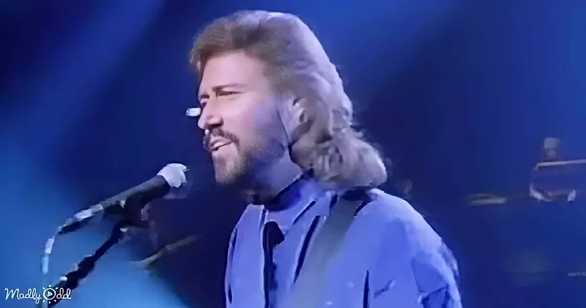 A man with long hair and a beard sings into a microphone on stage, wearing a blue shirt and tie.