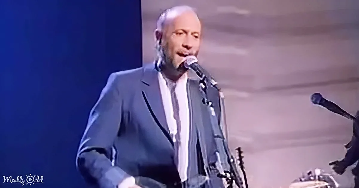 A man in a suit sings into a microphone on stage, with a guitar visible in the background.