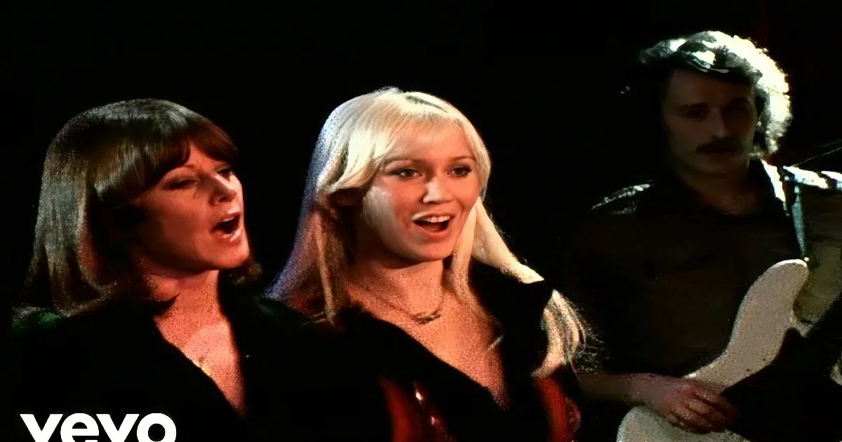 A woman with long blonde hair plays guitar on stage, singing, with a drummer and other musicians in the background.