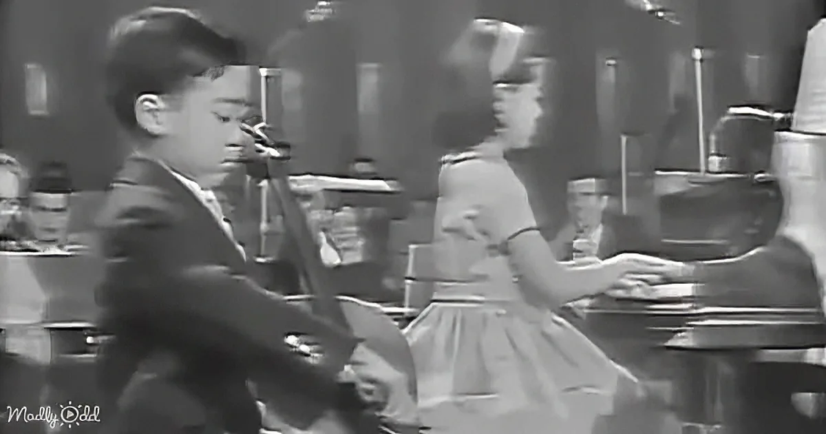 A boy in a suit dances with a girl in a skirt, both mid-movement, in a lively setting.