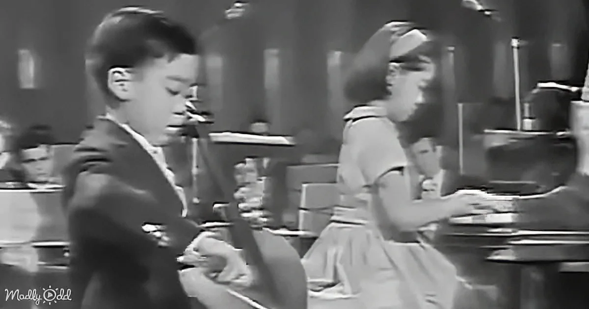 A boy in a suit dances while a girl in a dress sits at a diner counter.