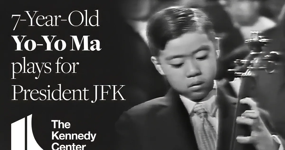 A young boy in a suit and tie looks down, holding a small object, with blurred figures in the background.