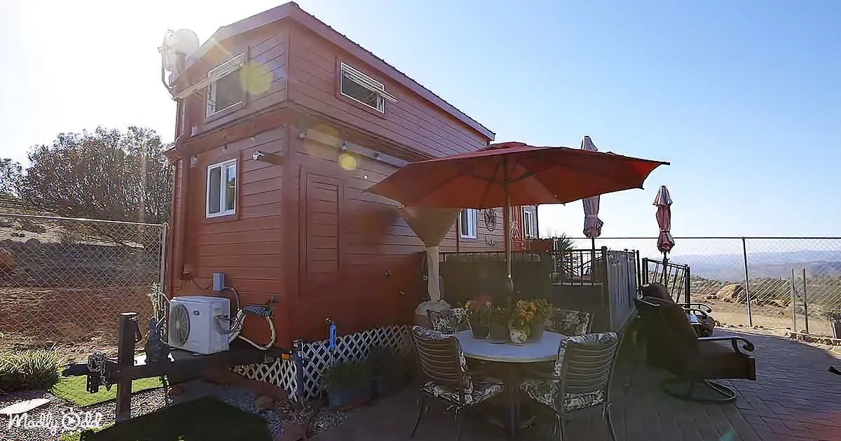 A red tiny house with a patio featuring a table, chairs, and a red umbrella under a clear sky.