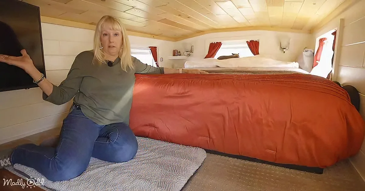 A woman kneels on a rug, gesturing towards a large, orange-covered object in a cozy, wooden interior space.