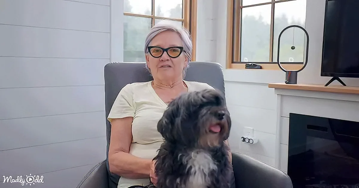 A woman sits in a chair with a small black dog on her lap, both looking at the camera.