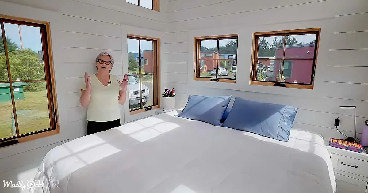 A woman stands in a bright, modern bedroom with white walls, large windows, and a neatly made bed with blue pillows.