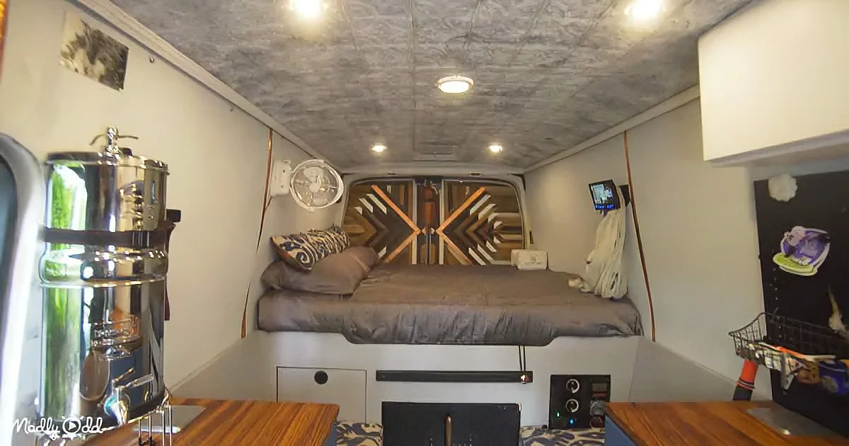 A compact, well-organized van interior with a bed, storage, and a large water tank.