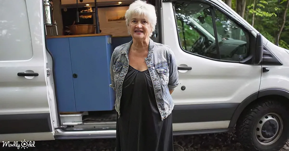 A woman stands in front of a van with an open door, revealing a kitchen setup inside.