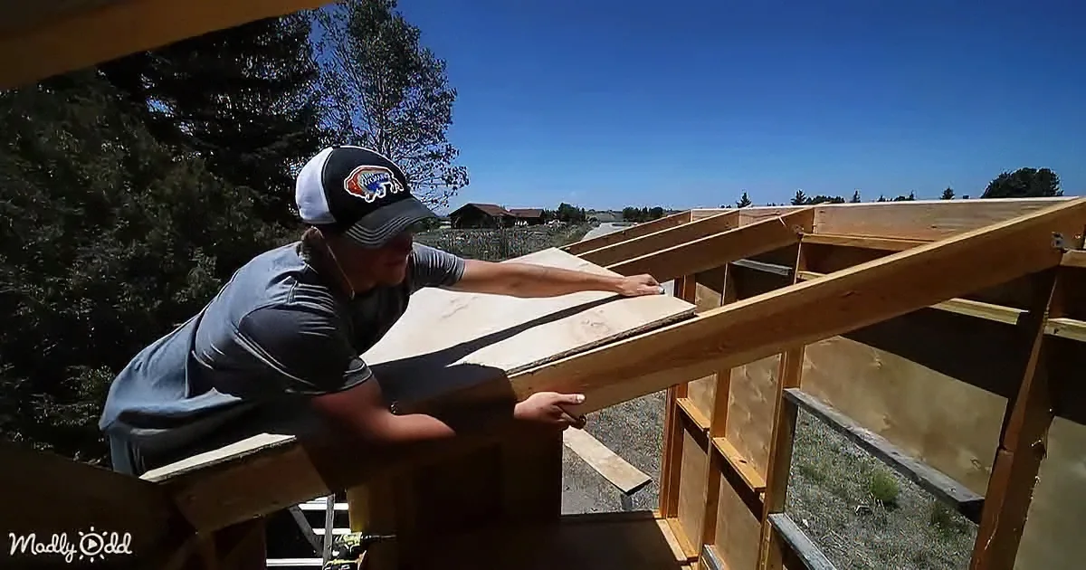 A man in a cap works on a wooden structure outdoors, surrounded by trees and a clear sky.
