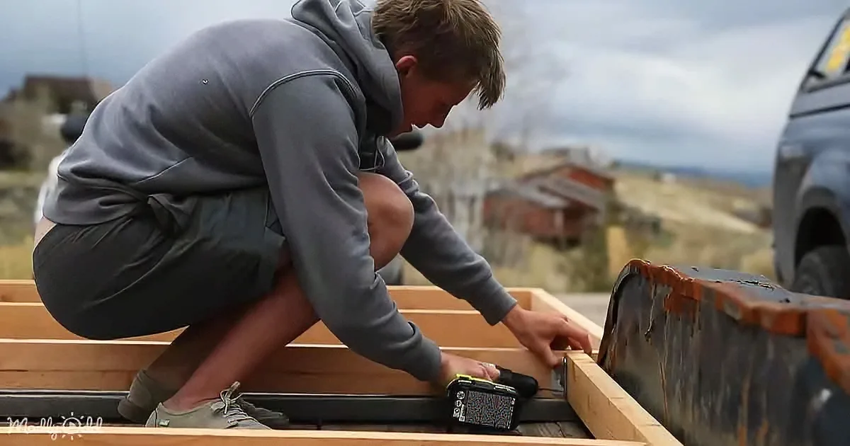 A person in a grey hoodie is crouching, working on a wooden structure with a power tool.