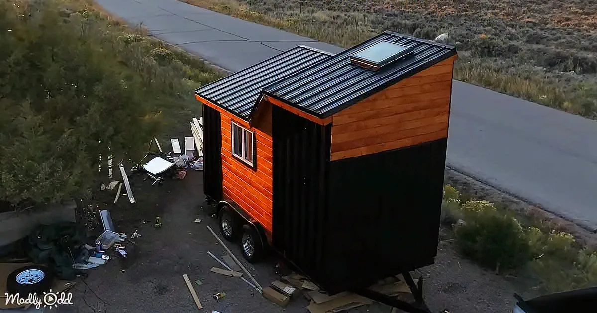 A tiny house with a wooden exterior and black roof sits on a trailer, surrounded by construction materials and a rural...