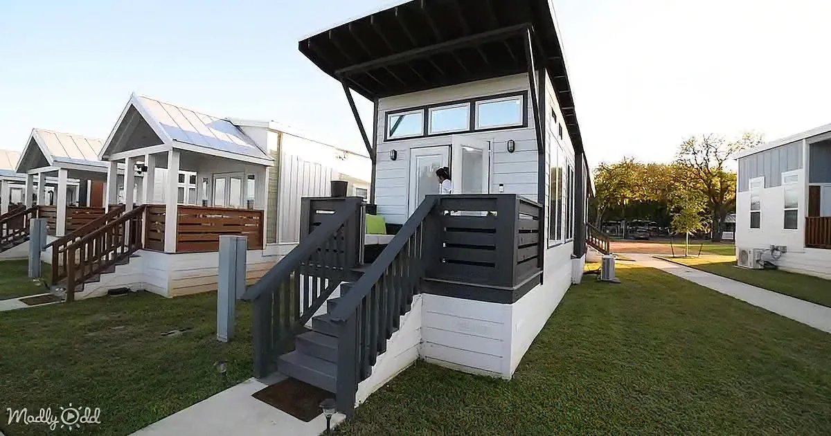 A modern, white and black tiny house with a wooden deck and stairs stands on a grassy lawn.