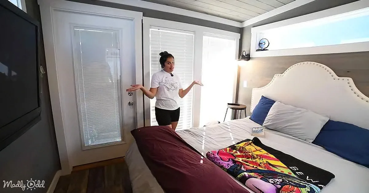 A woman stands in a cozy bedroom with a bed, colorful blanket, and a door with blinds.