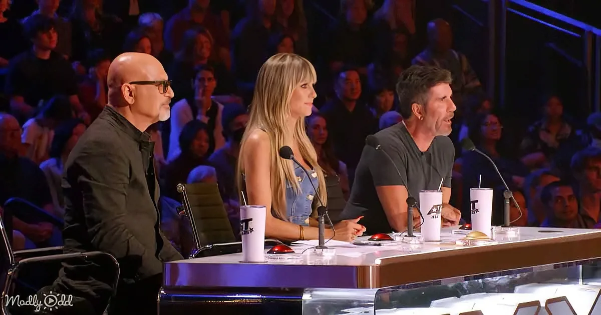 Three judges sit at a table with microphones and cups, facing an audience in a dimly lit room.