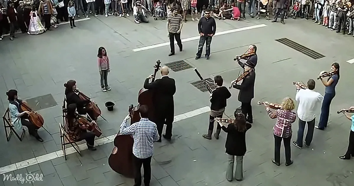 A street orchestra performs in a square, surrounded by an audience, with musicians playing string instruments.