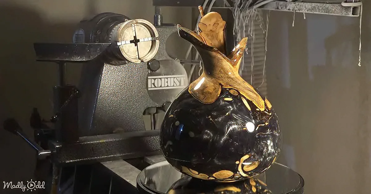 A glossy black vase with gold accents sits on a stand in front of a lathe machine.