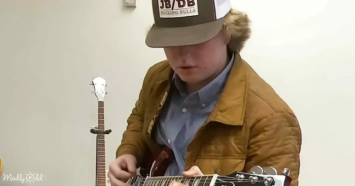 A person wearing a brown jacket and a cap is playing a guitar.