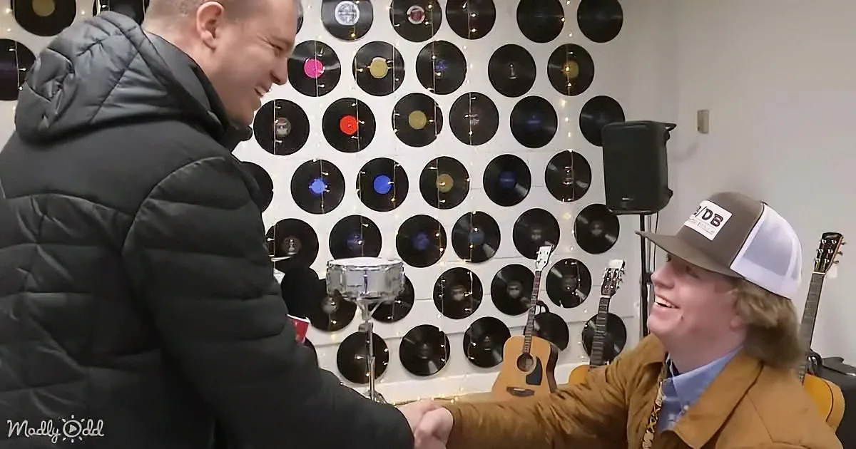 Two men in a room with vinyl records on the wall, one in a black jacket smiling, the other in a brown jacket and hat...