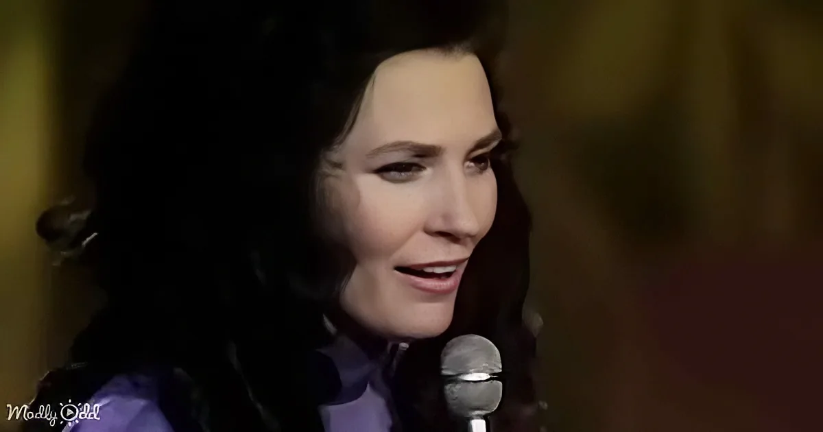 A woman with dark hair and a purple top sings into a microphone, her eyes closed.