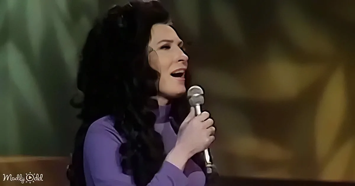 A woman with long dark hair sings into a microphone, wearing a purple top, against a blurred green and yellow background.
