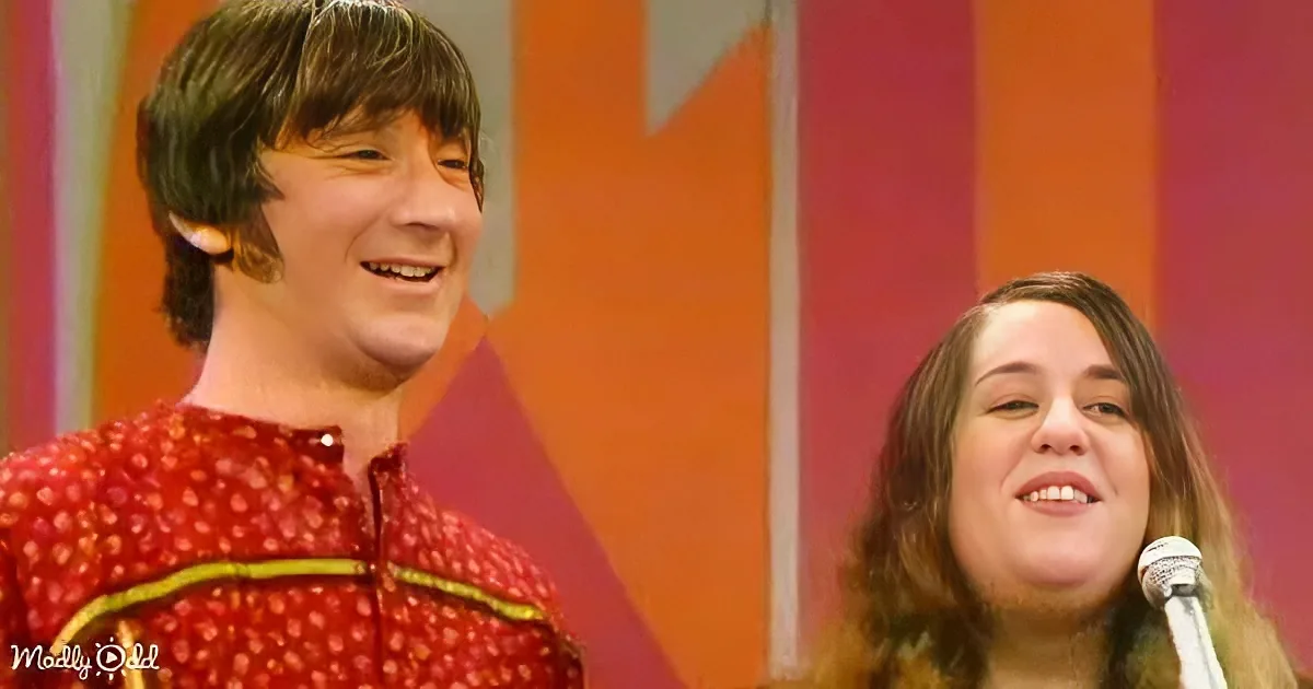 A man in a red patterned shirt smiles beside a woman with a microphone, both against a colorful striped background.