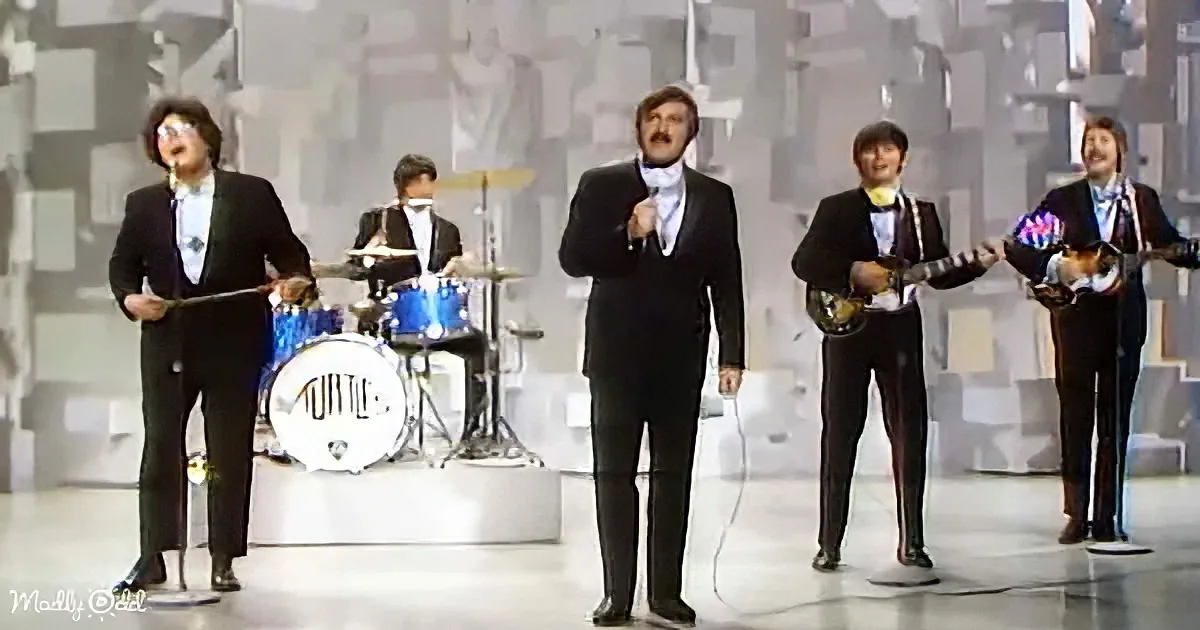 A band performing on stage, dressed in black suits, with a drummer, a lead singer, and two guitarists.
