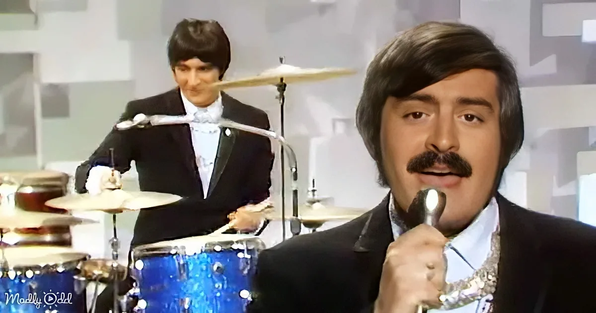 A man in a suit sings into a microphone while another man plays drums in the background.