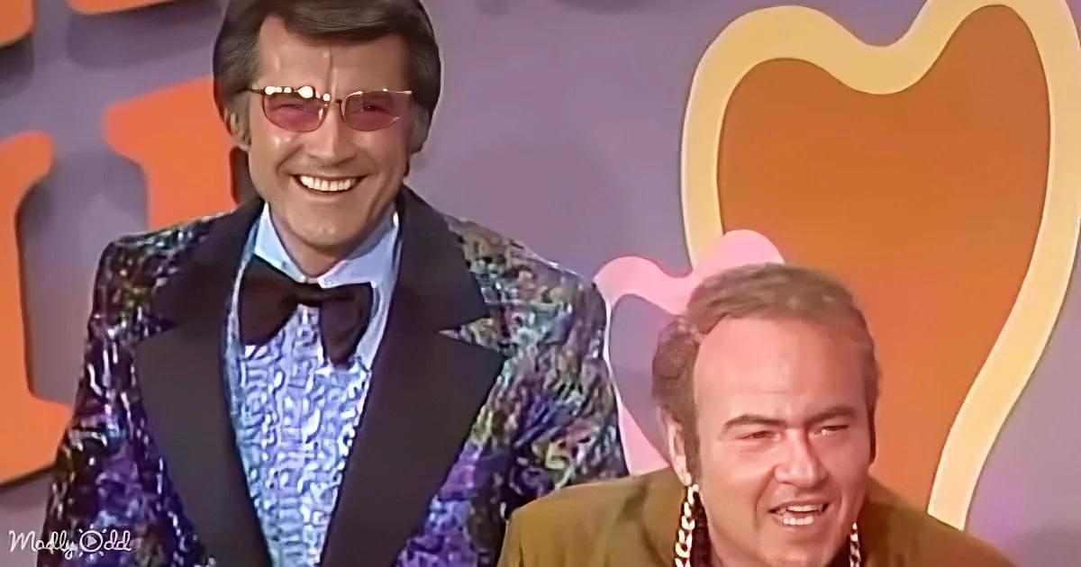 A man in a shiny, patterned suit and sunglasses smiles widely, while another man in a brown suit looks down, both against...