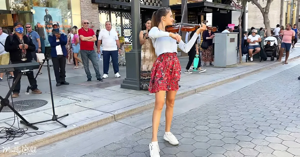 A woman plays violin on sidewalk, surrounded by onlookers and street performers.