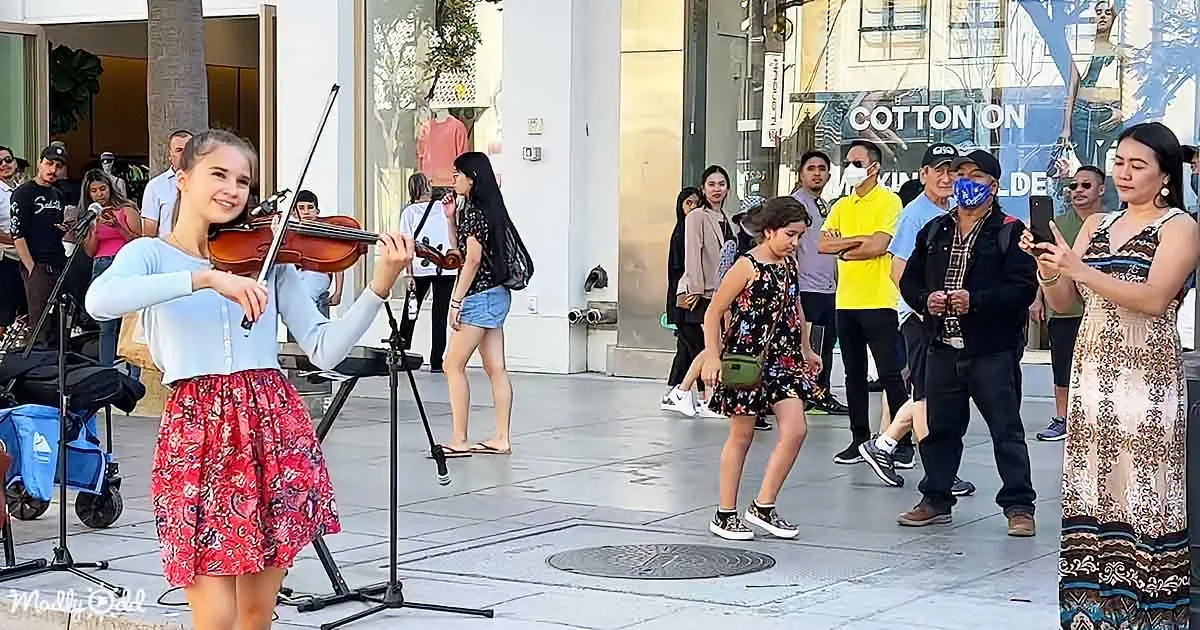A young girl plays violin on a city street, surrounded by onlookers and passersby.