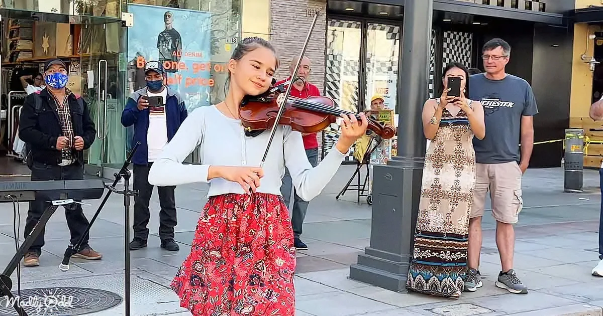 A young woman in a floral skirt plays the violin on a city street, surrounded by onlookers and a musician.