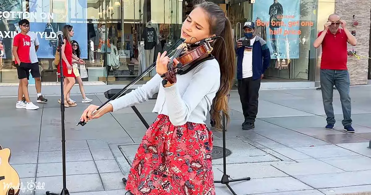 A young woman plays violin on a city street, surrounded by pedestrians and storefronts.