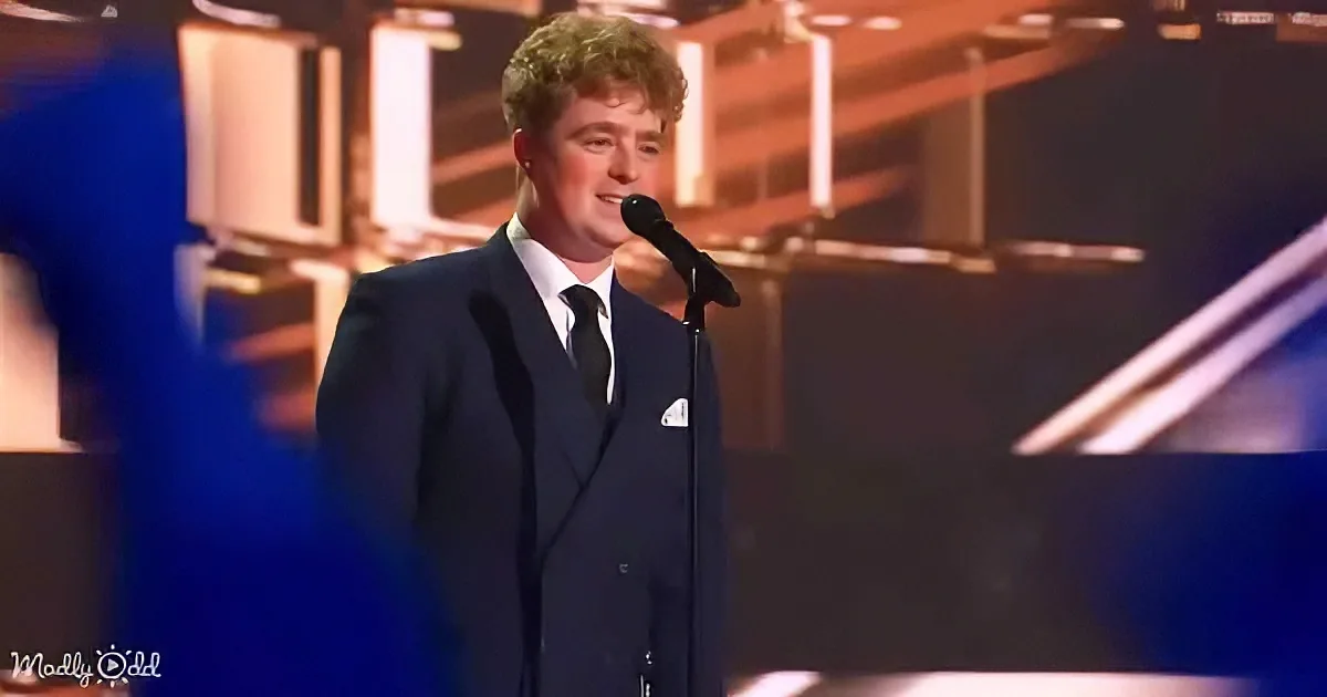 A man in a suit stands on stage, holding a microphone, with a spotlight on him.
