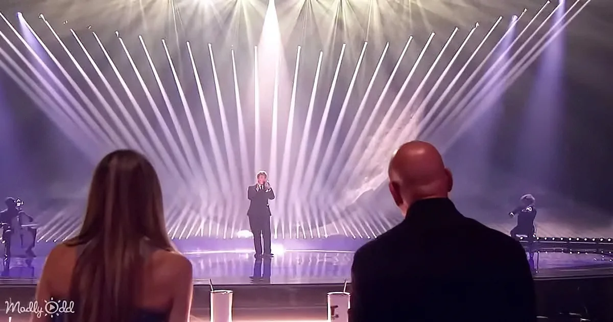 A person stands on a brightly lit stage with beams of light, surrounded by judges and a band.