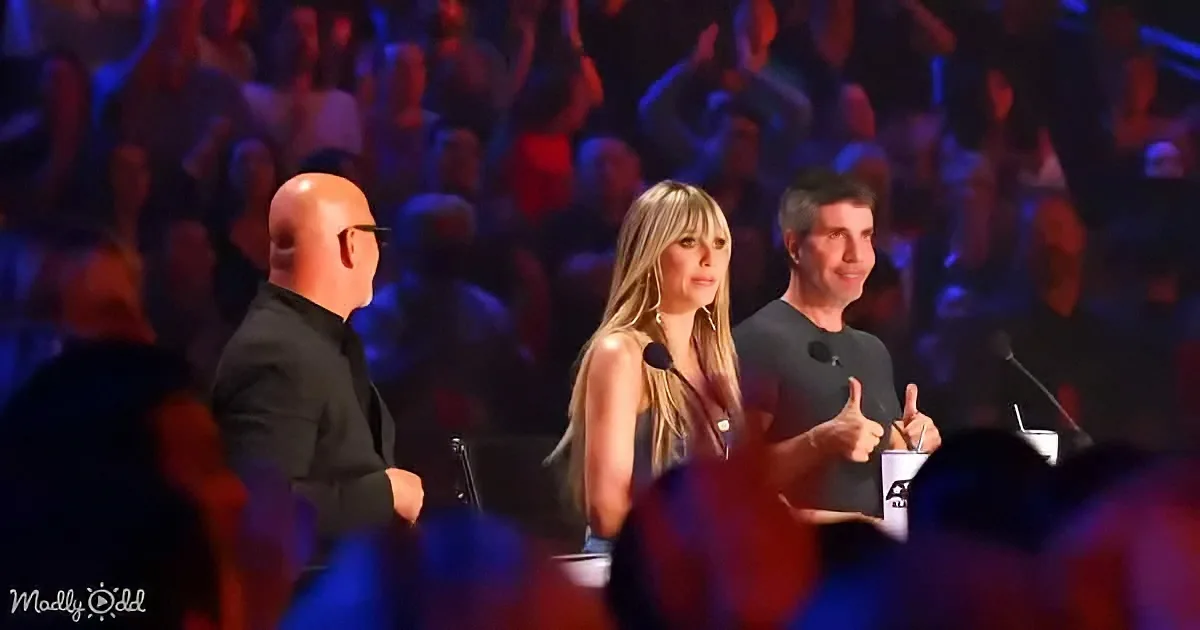 A woman with long blonde hair stands on stage with two men, one giving a thumbs-up, in front of a large audience.