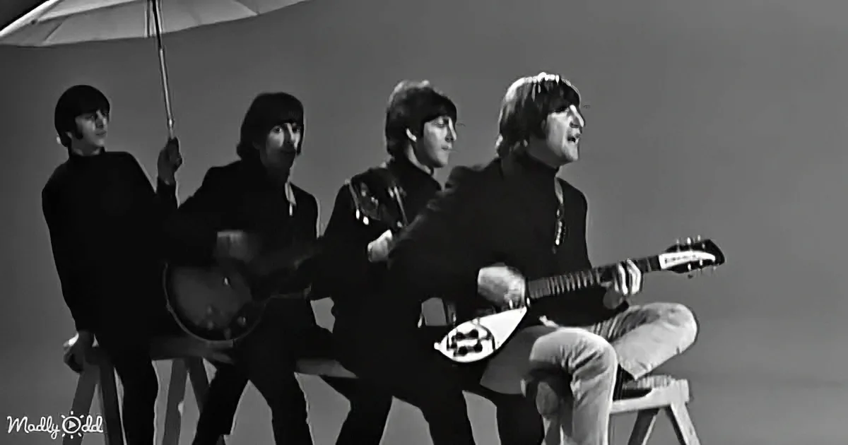 The Beatles play guitars on stools, one holding an umbrella, in a black-and-white photo.