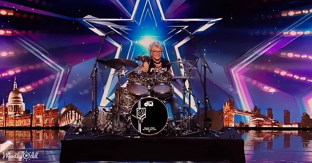 A drummer sits behind a drum set on a stage with a cityscape and a large star backdrop.