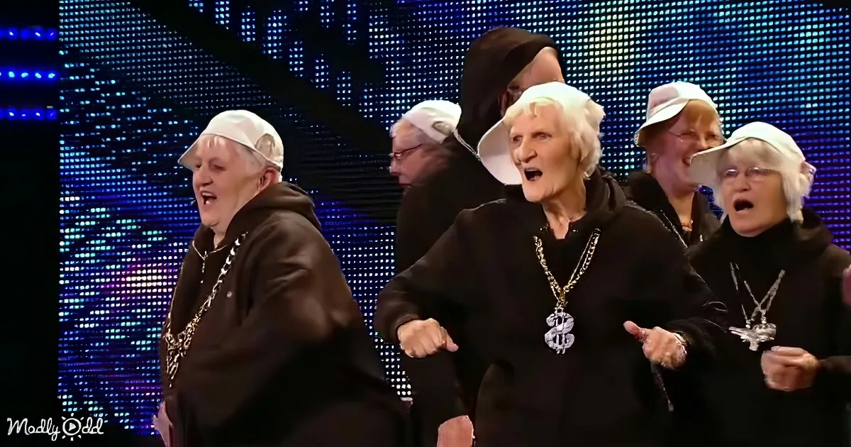 A group of elderly women in black outfits and white hats, adorned with dollar sign necklaces, are dancing and laughing on...