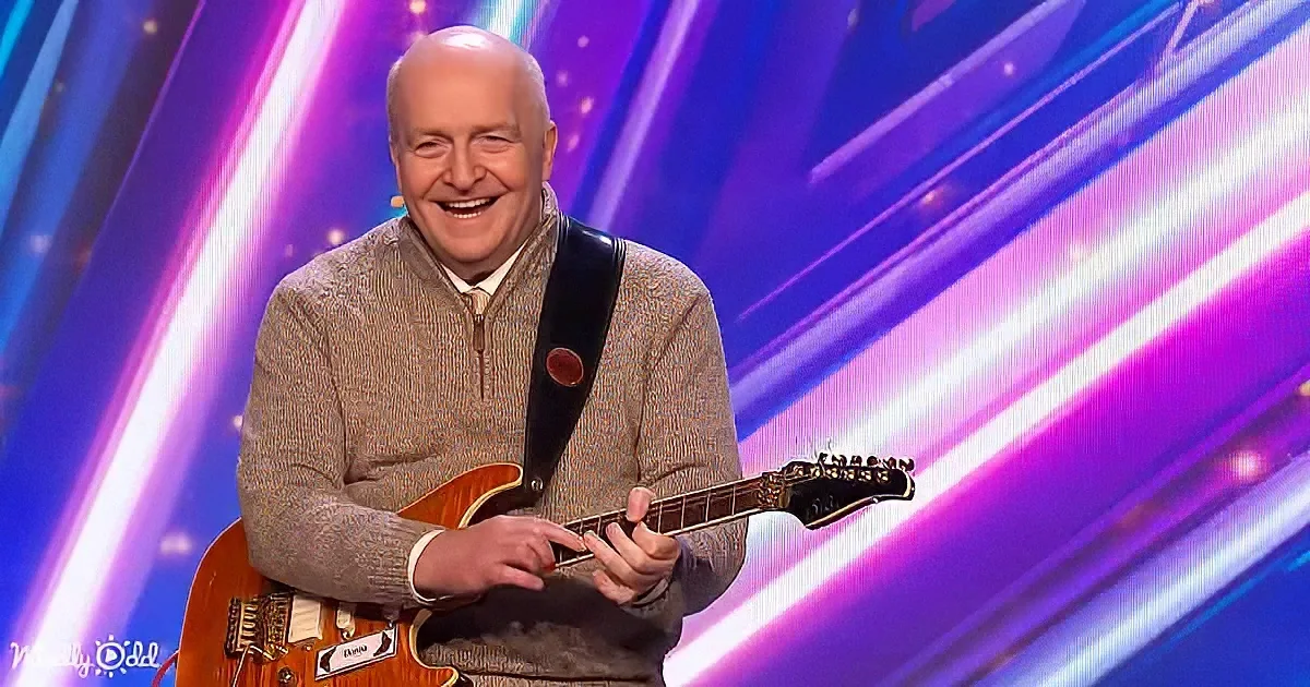 A man in a sweater smiles while playing a guitar on a stage with colorful lights.