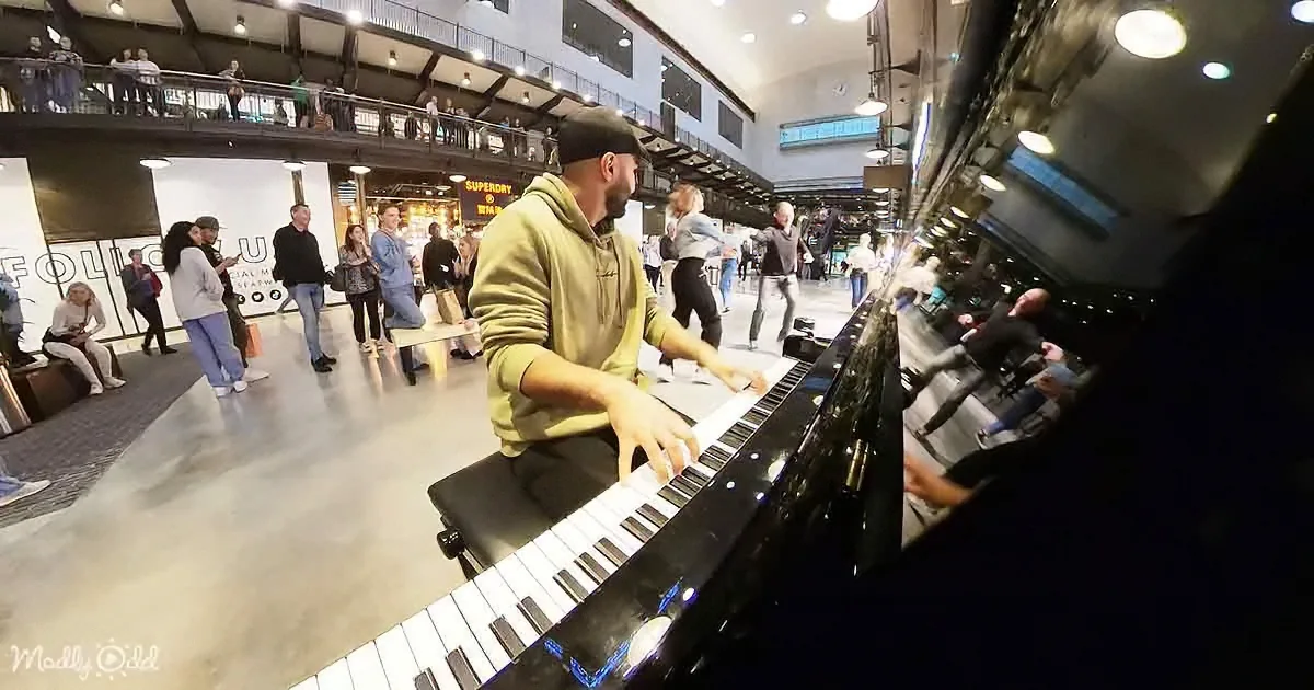 A man in a green hoodie plays a piano in a bustling indoor space with people walking around.