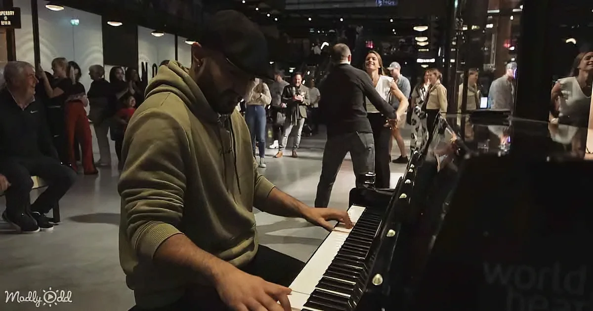A man in a hoodie plays a piano in a crowded, well-lit indoor space.
