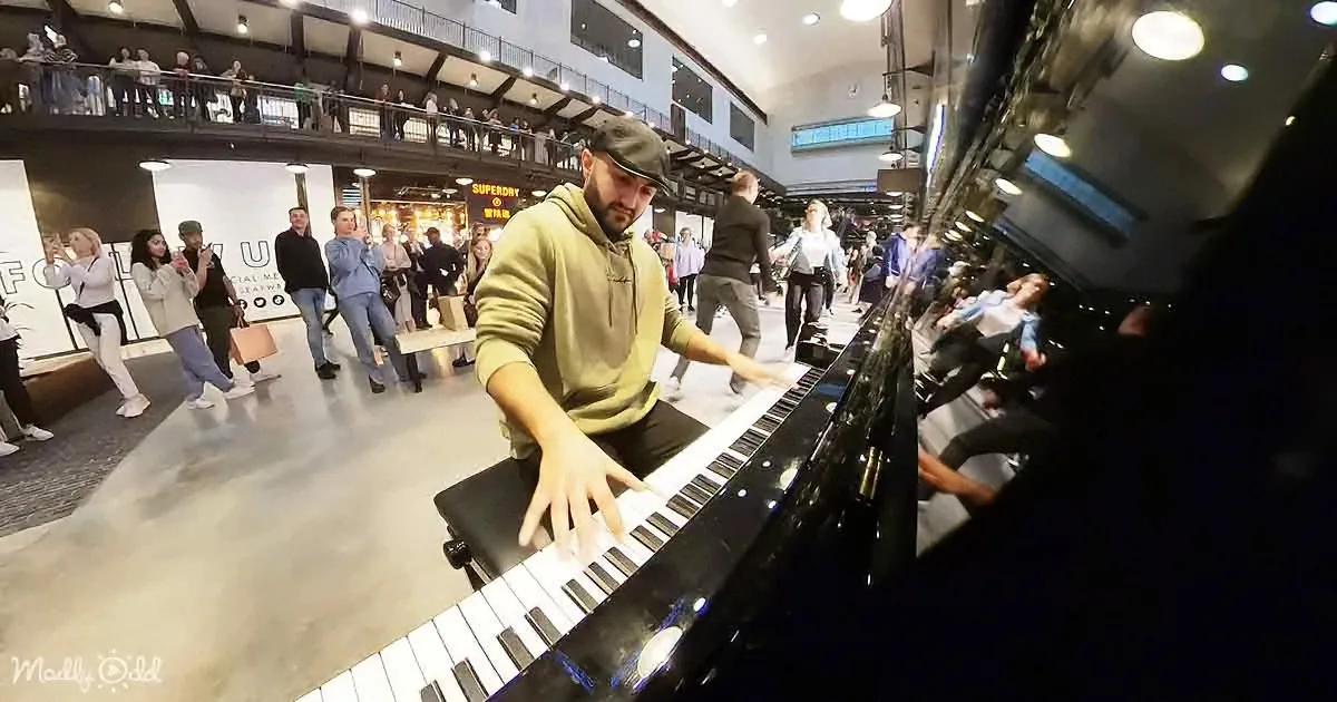 A man plays a piano in a busy, modern indoor space with people watching and taking photos.