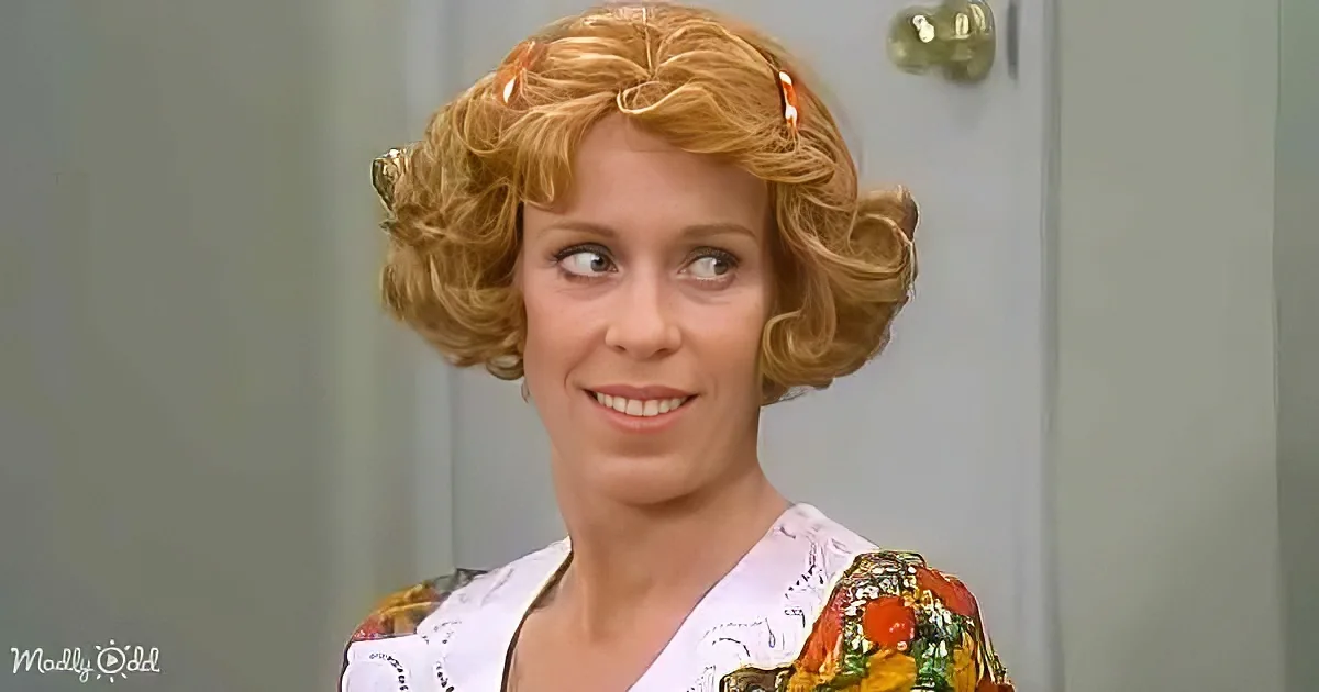 A woman with blonde, curly hair styled with clips, wearing a white blouse with colorful floral sleeves, smiles while...