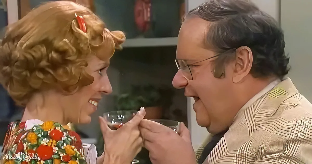 A woman with blonde hair and a floral dress holds a small glass, smiling at a man in a striped suit who is about to drink...