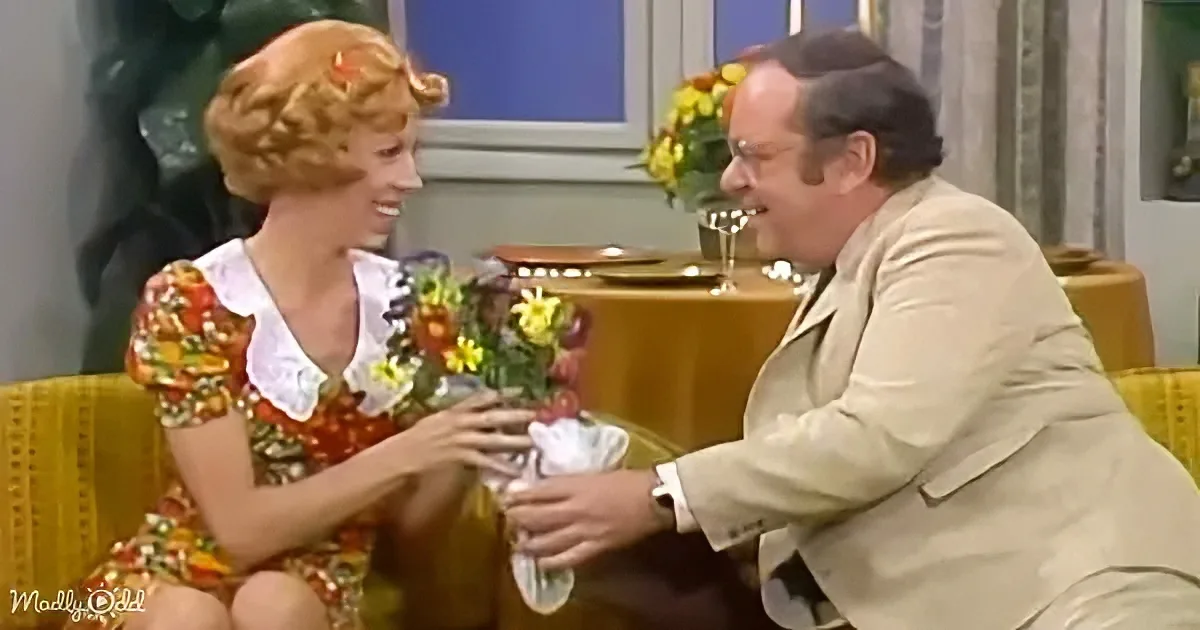 A woman in a floral dress hands a bouquet to a man in a tan suit, both smiling.