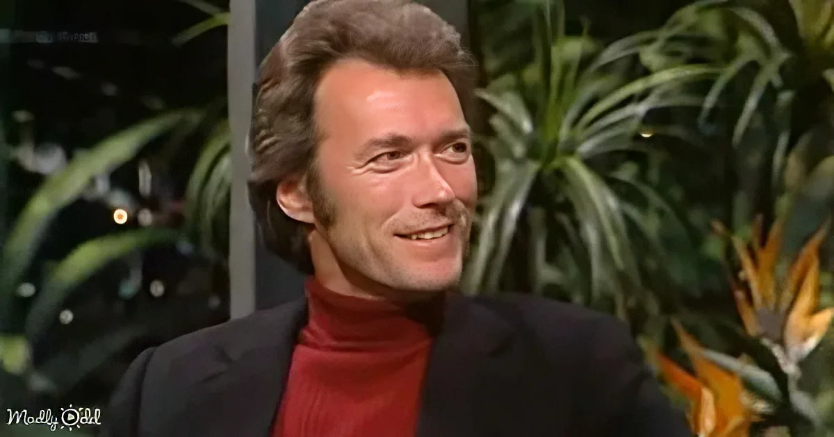 A man in a black suit and red turtleneck smiles, with greenery and a plant in the background.