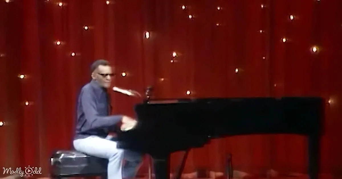 A man in glasses sits at a grand piano, wearing a dark jacket and light pants, against a red backdrop with string lights.
