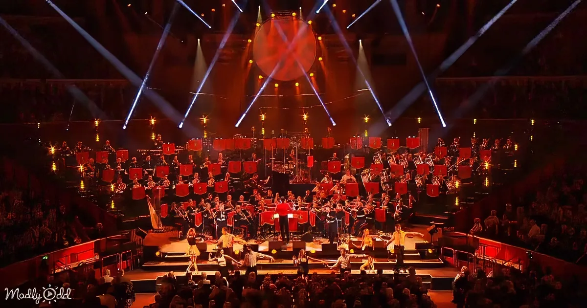A large orchestra with red instruments and performers on stage in front of a large audience.