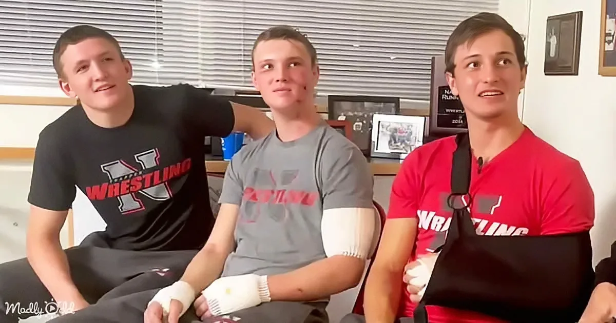 Three young wrestlers in red and gray shirts sit together, one with a cast on his arm, in a room with blinds and framed...
