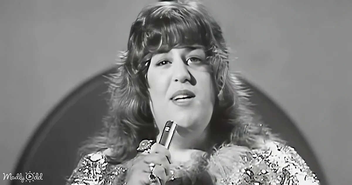 A person with long, wavy hair and a sparkly outfit is holding a microphone.